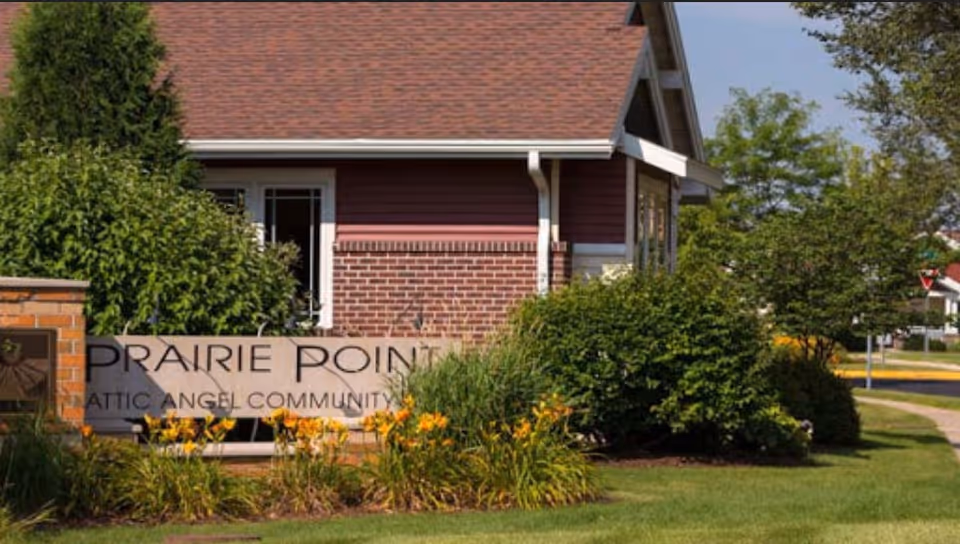 Exterior view of a building with red siding and a brown shingled roof, surrounded by green bushes and yellow flowers. In front of the building is a stone sign that reads 'Prairie Point Attic Angel Community'.