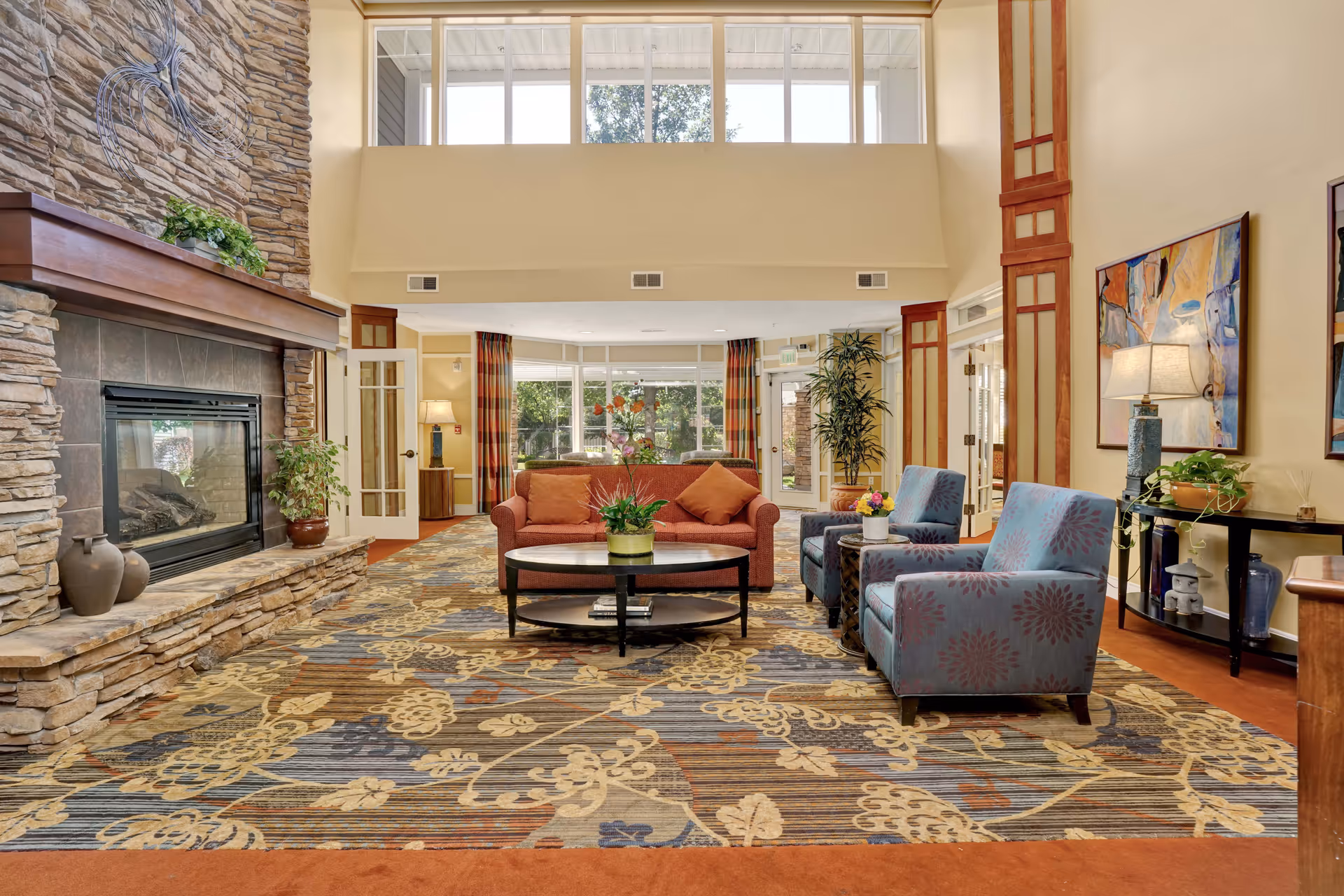 A spacious and well-lit living room area with a large stone fireplace on the left, a patterned carpet, a red sofa with orange cushions, two blue armchairs with floral patterns, a round coffee table with a plant, and large windows letting in natural light.