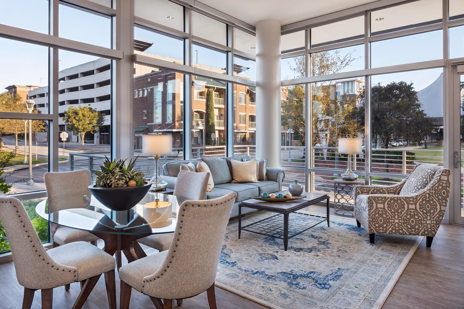 A bright and modern living area with large floor-to-ceiling windows showing an urban street view. The room features a glass round dining table with four upholstered chairs, a light blue sofa with cushions, a patterned armchair, a wooden coffee table with decorative items, two table lamps, and a blue and beige patterned rug on wooden flooring.