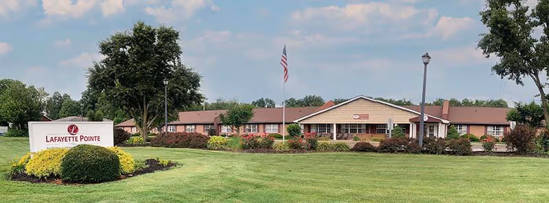 Exterior view of Lafayette Pointe senior living facility with a large green lawn, trees, and a sign displaying the facility name. The building is a single-story structure with a brown roof and beige walls, and an American flag is flying on a flagpole in front.