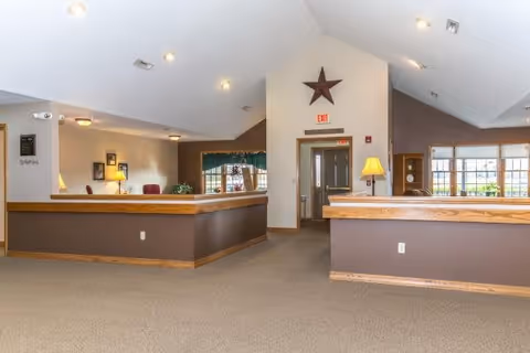 Interior view of a senior living facility reception area with a high vaulted ceiling, beige carpet, brown walls with wooden trim, two reception desks with lamps, a large star decoration on the wall above an exit door, and windows letting in natural light.