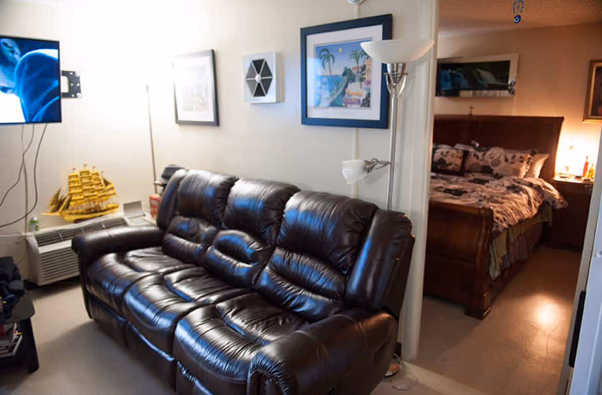 Interior view of a living area with a dark brown leather couch, a floor lamp, framed artwork on the wall, and a mounted TV. Through an open doorway, a bedroom is visible with a wooden bed frame, patterned bedding, and a nightstand with a lamp.