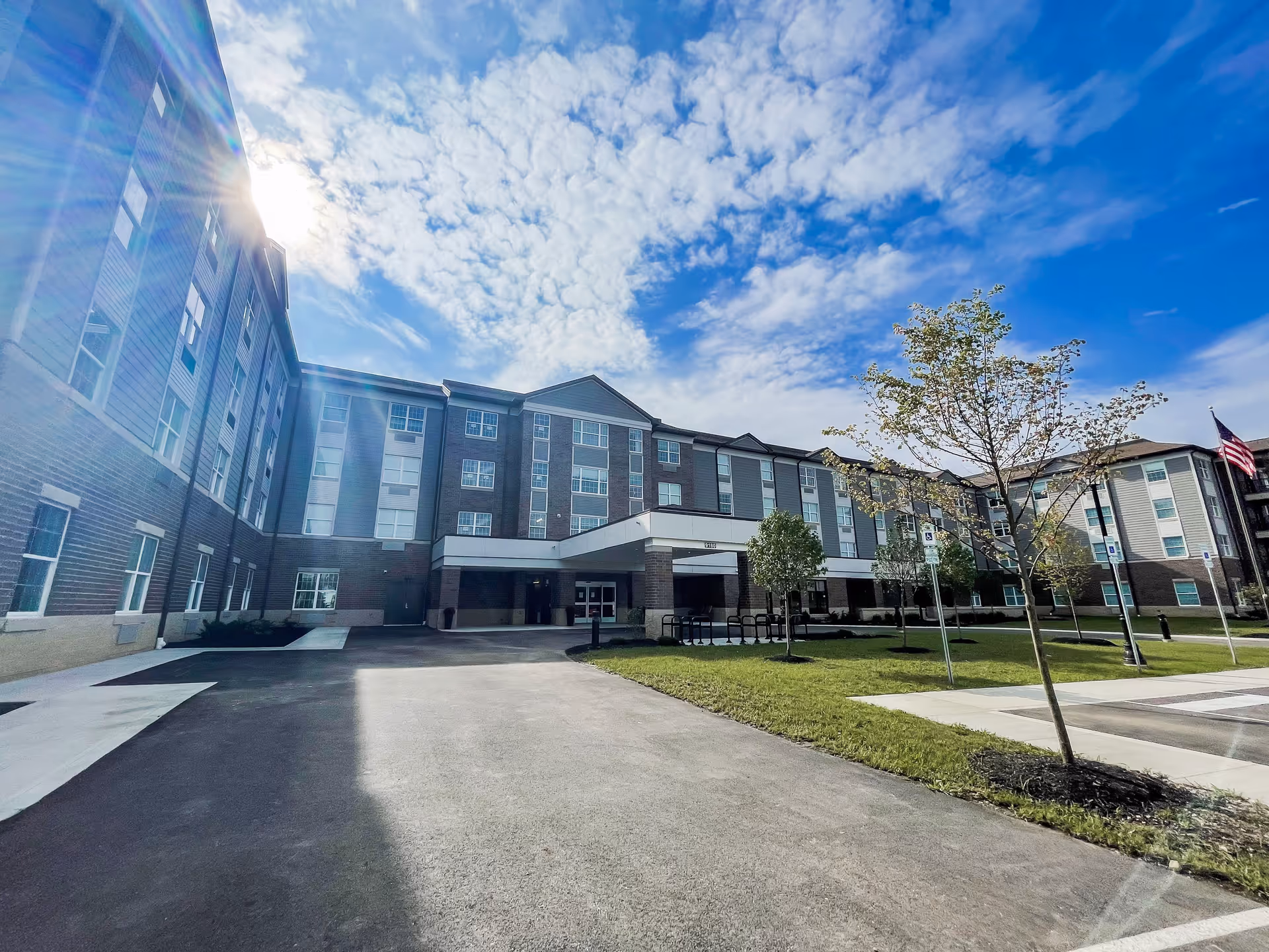 Exterior view of a multi-story senior living facility building under a partly cloudy blue sky with the sun shining from the left. The building has multiple windows, a covered entrance, and a driveway leading up to it. There are small trees and a grassy area in front of the building along with a few parking spaces.