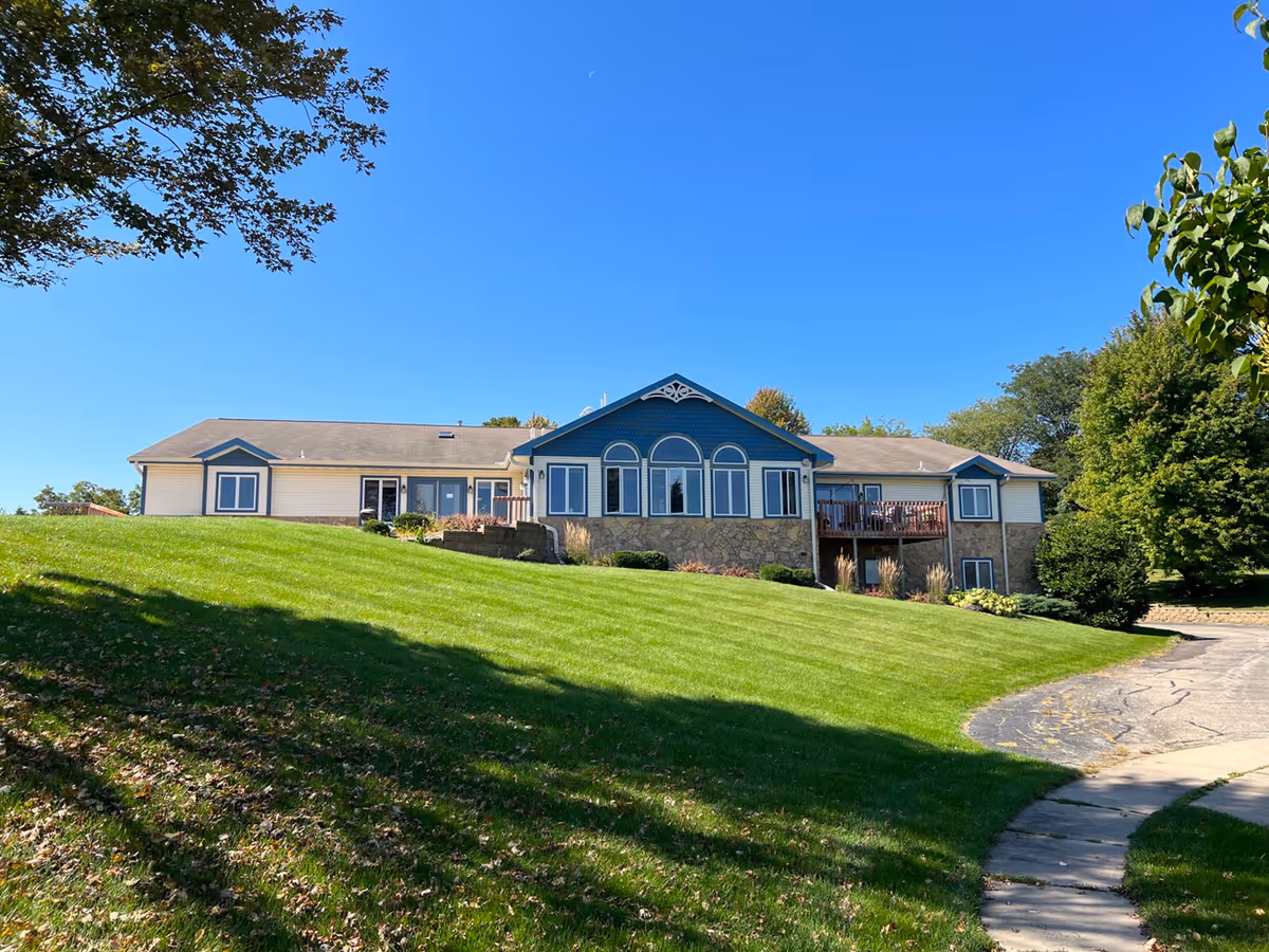 Exterior view of a single-story residential building with beige and blue siding, stone accents, and multiple windows. The building is situated on a well-maintained grassy hill with a curved driveway leading up to it. Trees and clear blue sky are visible in the background.