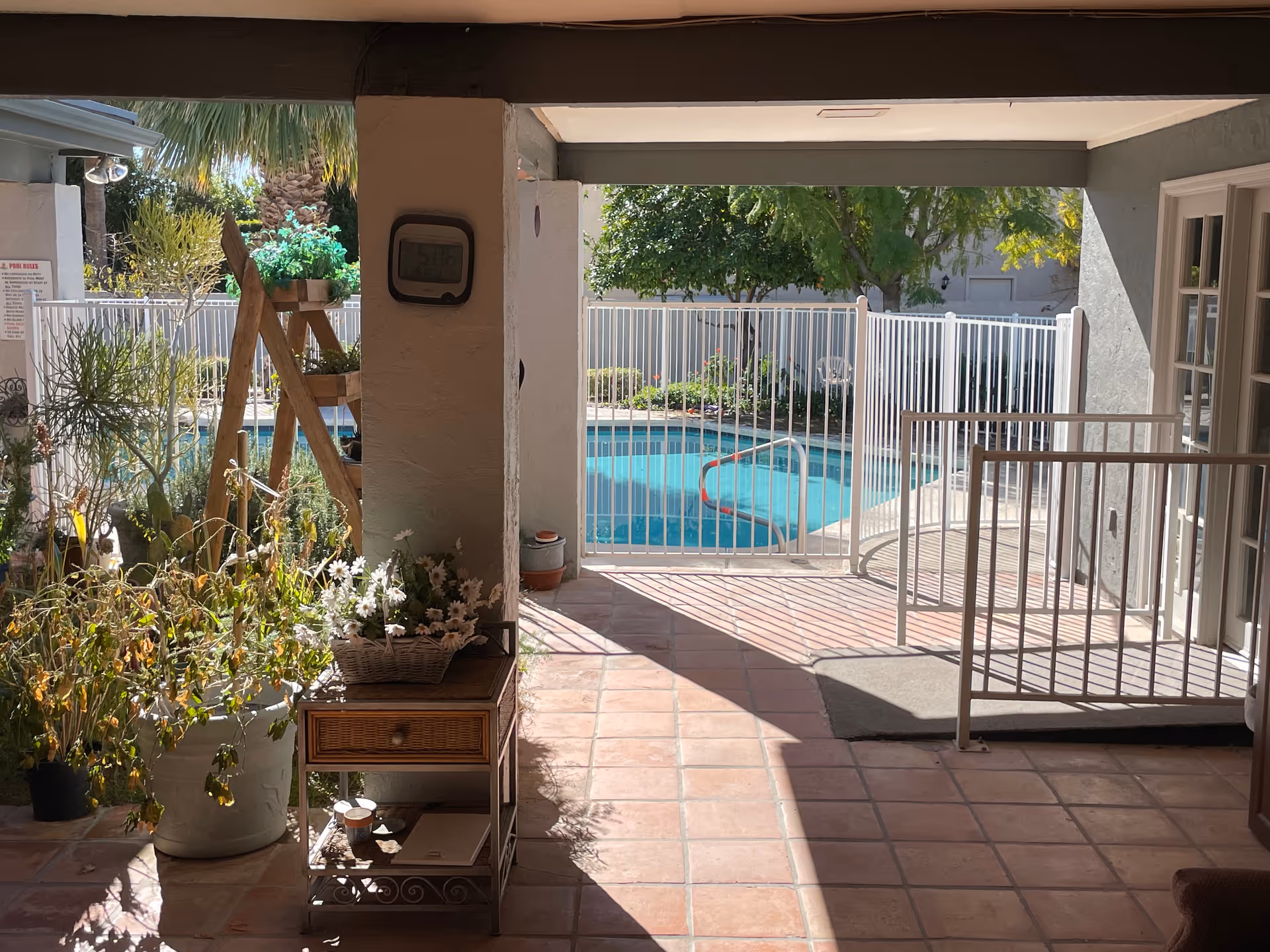 View of a covered patio area with potted plants and a small table with flowers, leading to a fenced swimming pool surrounded by greenery and trees.
