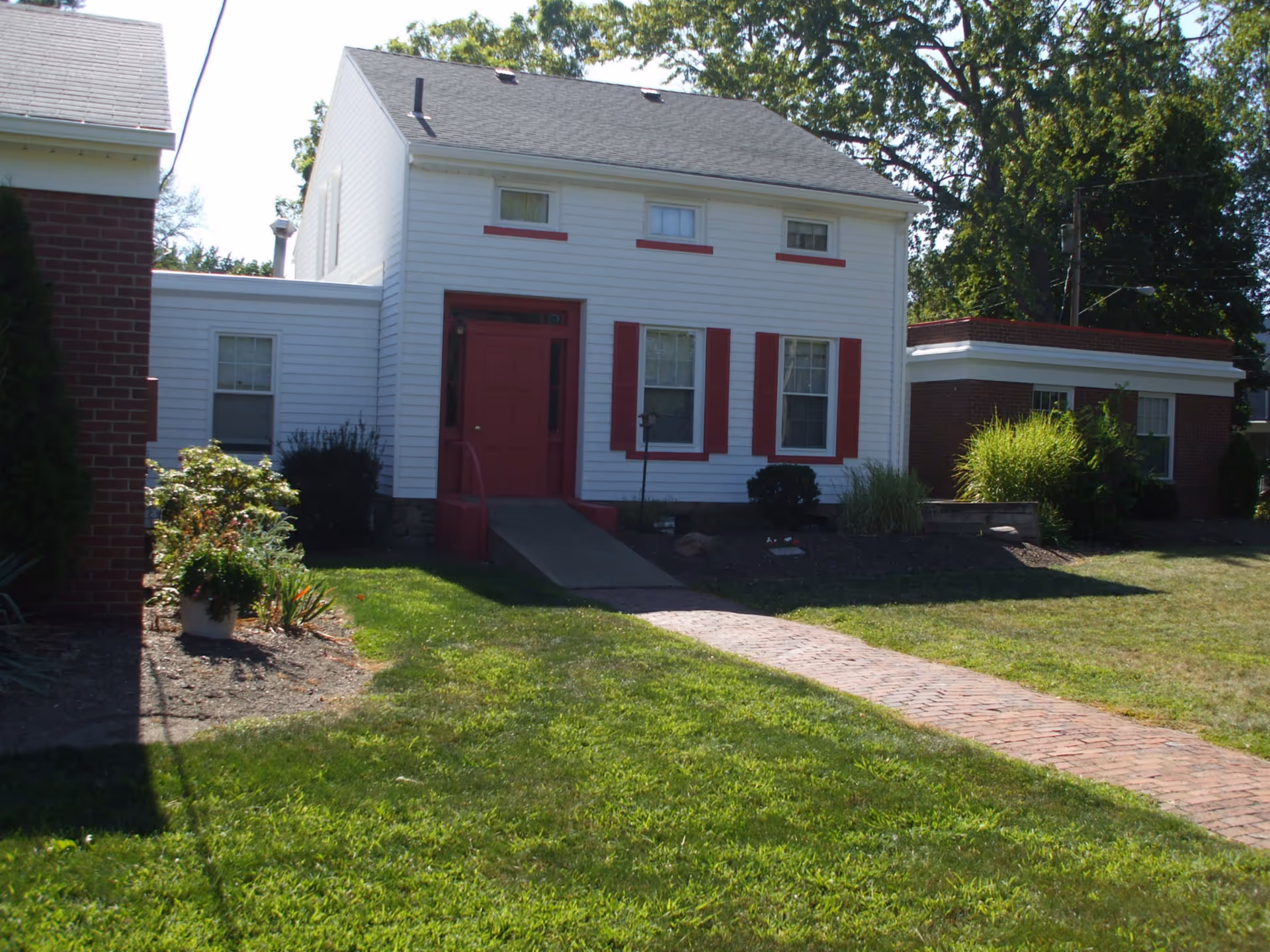Exterior view of a white two-story house with red trim and a red front door, surrounded by green grass and some bushes. A brick pathway leads up to the front entrance. There is a smaller brick building to the right and part of another building to the left.