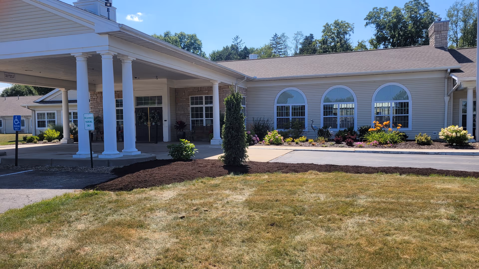 Exterior view of Whispering Pines Village senior living facility showing the entrance with white columns, a covered drop-off area, landscaped garden beds with flowers, and a grassy lawn in the foreground under a clear blue sky.