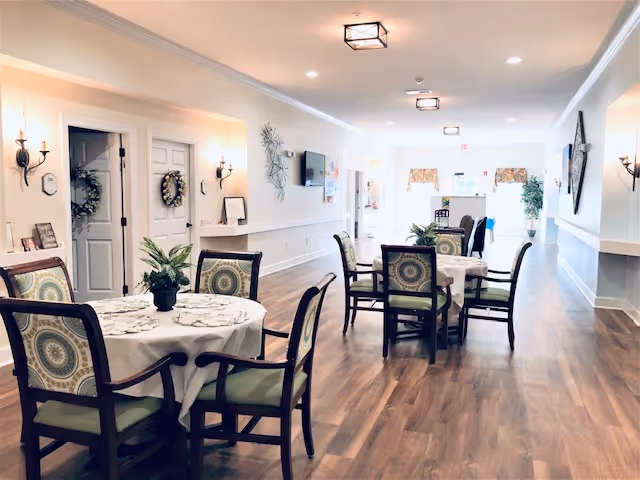 A bright and spacious dining area with round tables covered in white tablecloths, each surrounded by wooden chairs with patterned cushions. The room has wooden flooring, wall sconces, decorative plants on the tables, and a large window at the far end letting in natural light.