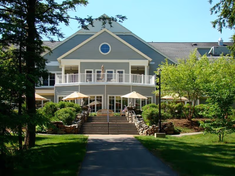 Exterior view of a two-story senior living facility building with a gray facade, white trim, and a balcony on the second floor. The entrance is accessed by a set of stairs flanked by stone walls and greenery. There are several patio umbrellas and outdoor seating areas surrounded by well-maintained bushes and trees.