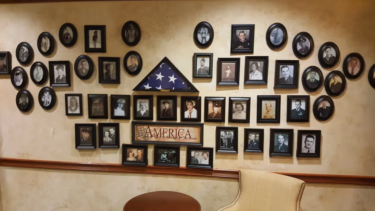 A wall decorated with numerous framed black and white and color photographs of military personnel, arranged around a folded American flag in a triangular display case and a framed sign that reads 'AMERICA'. Below the photos is a wooden chair rail and part of a beige upholstered chair and a round wooden table.