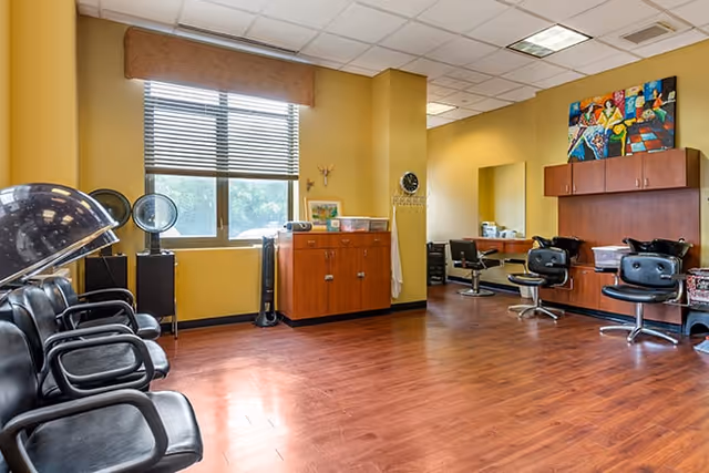 Interior of a salon area in a senior living facility with black salon chairs, hair dryers, wooden cabinets, a large window with blinds, and colorful artwork on the wall.