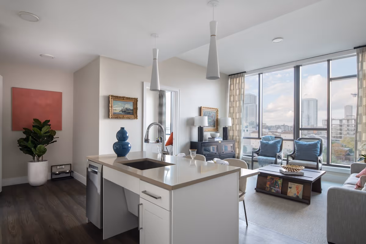A modern senior living apartment interior featuring a kitchen island with a sink and blue decorative vase, a living area with two armchairs with blue cushions, a coffee table with books and a decorative bowl, a gray sofa, large floor-to-ceiling windows with a cityscape view, framed paintings on the walls, and a potted plant in the corner.