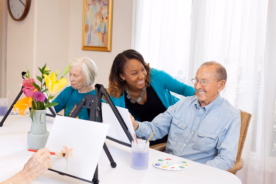 Two elderly individuals seated at a table painting on canvases with easels, accompanied by a smiling caregiver or instructor in a bright room with a vase of colorful flowers and a painting on the wall.