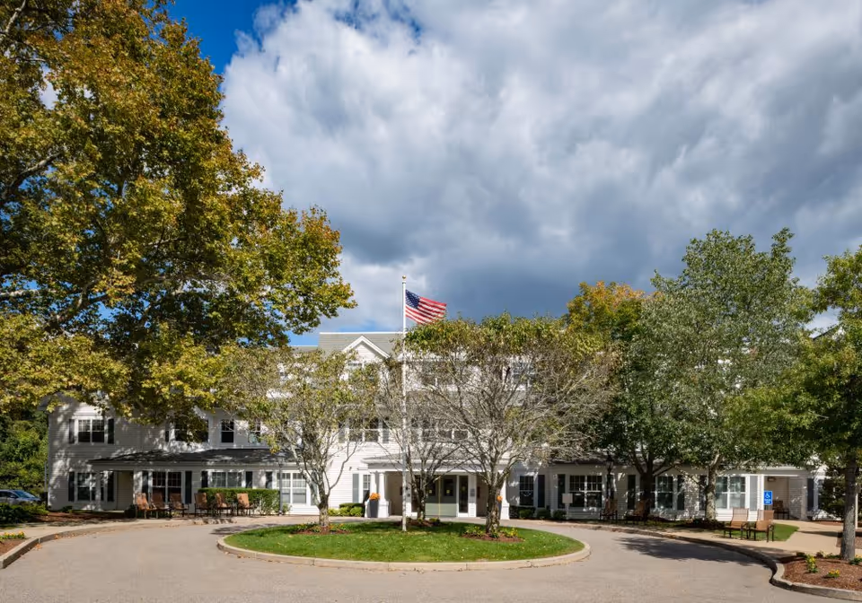 Front exterior view of a large white senior living facility building with multiple windows, surrounded by trees and greenery. An American flag is flying on a flagpole in front of the building under a partly cloudy sky. There is a circular driveway with benches and a wheelchair accessible parking sign visible.