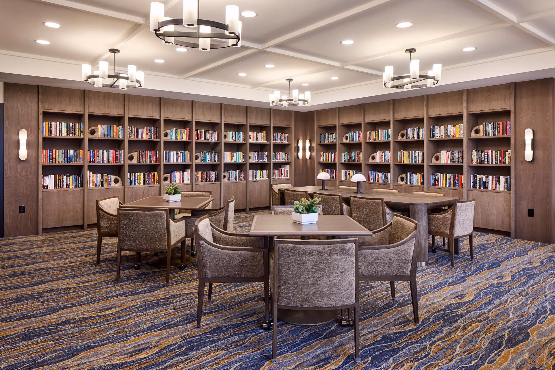 A spacious and well-lit library room with multiple wooden bookshelves filled with books along the walls. The room features several square tables surrounded by upholstered chairs, with small potted plants on the tables. Modern ceiling lights and wall sconces provide warm lighting, and the floor is covered with a patterned carpet in shades of blue and beige.