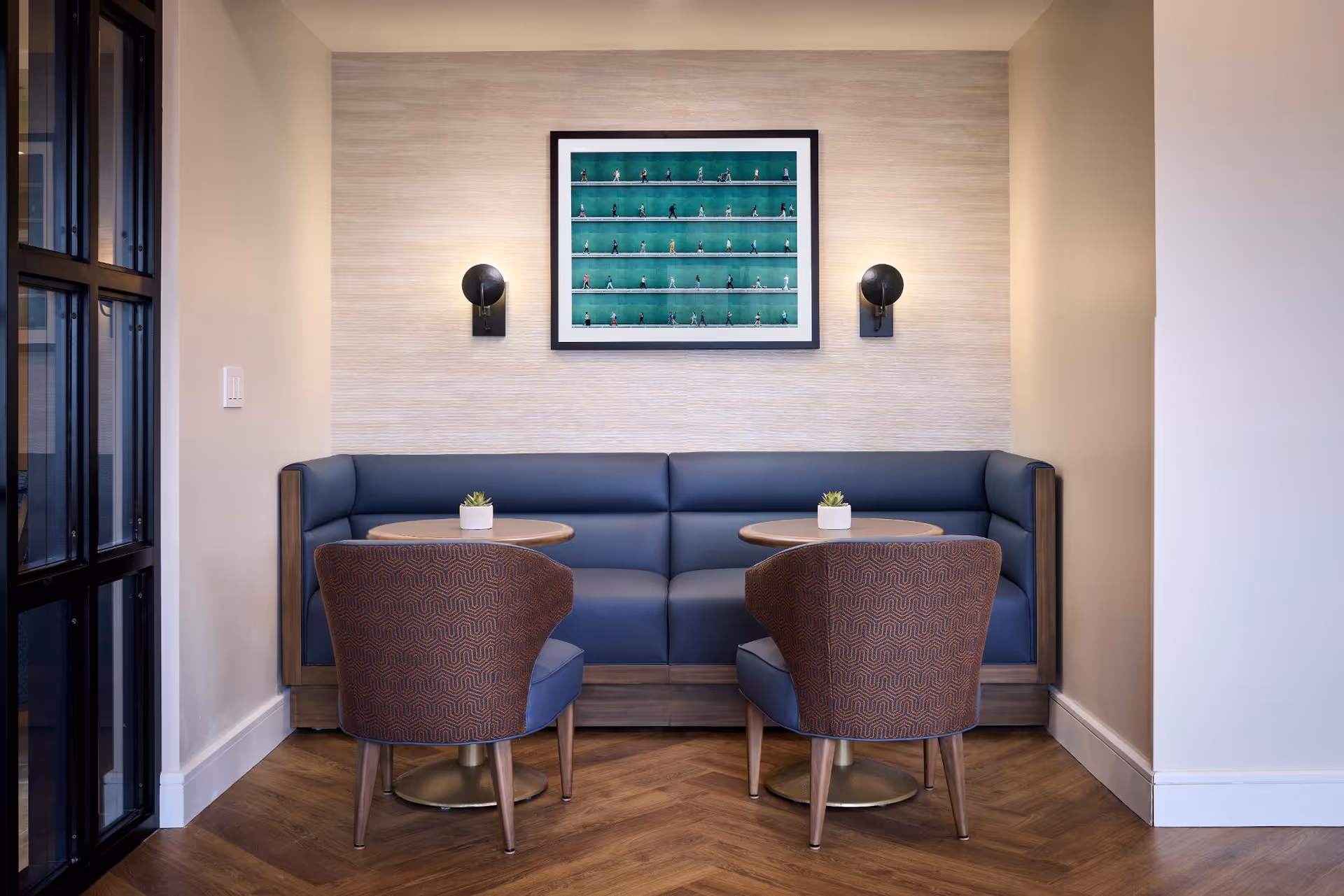A cozy seating area with a blue cushioned bench along the wall, two round wooden tables, and two patterned armchairs. The wall behind the bench has a framed artwork featuring multiple small figurines on green shelves and two black wall sconces providing light. The floor has a herringbone wood pattern.