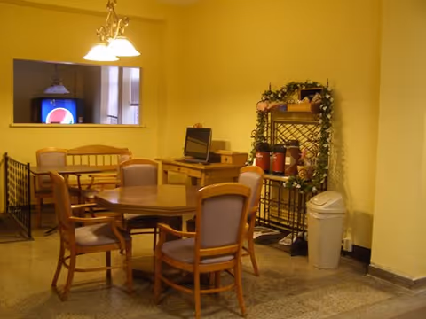 Interior room with a round wooden table surrounded by five chairs with cushions. In the background, there is a wooden bench and a small desk with a computer monitor. A decorative metal rack with plants and fire extinguishers is against the wall next to a white trash bin. The walls are painted yellow and the floor is tiled.