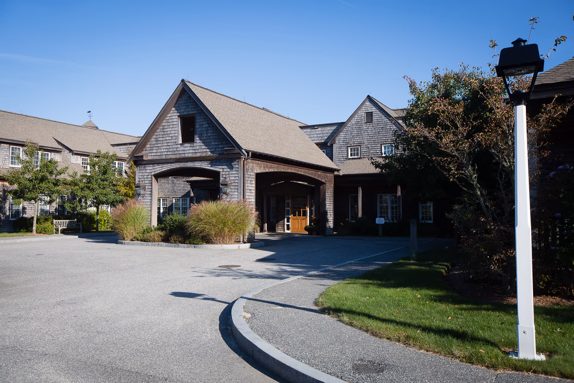 Exterior view of a senior living facility with a large covered entrance, wooden shingles on the building, landscaped bushes, trees, and a lamp post on a sunny day.