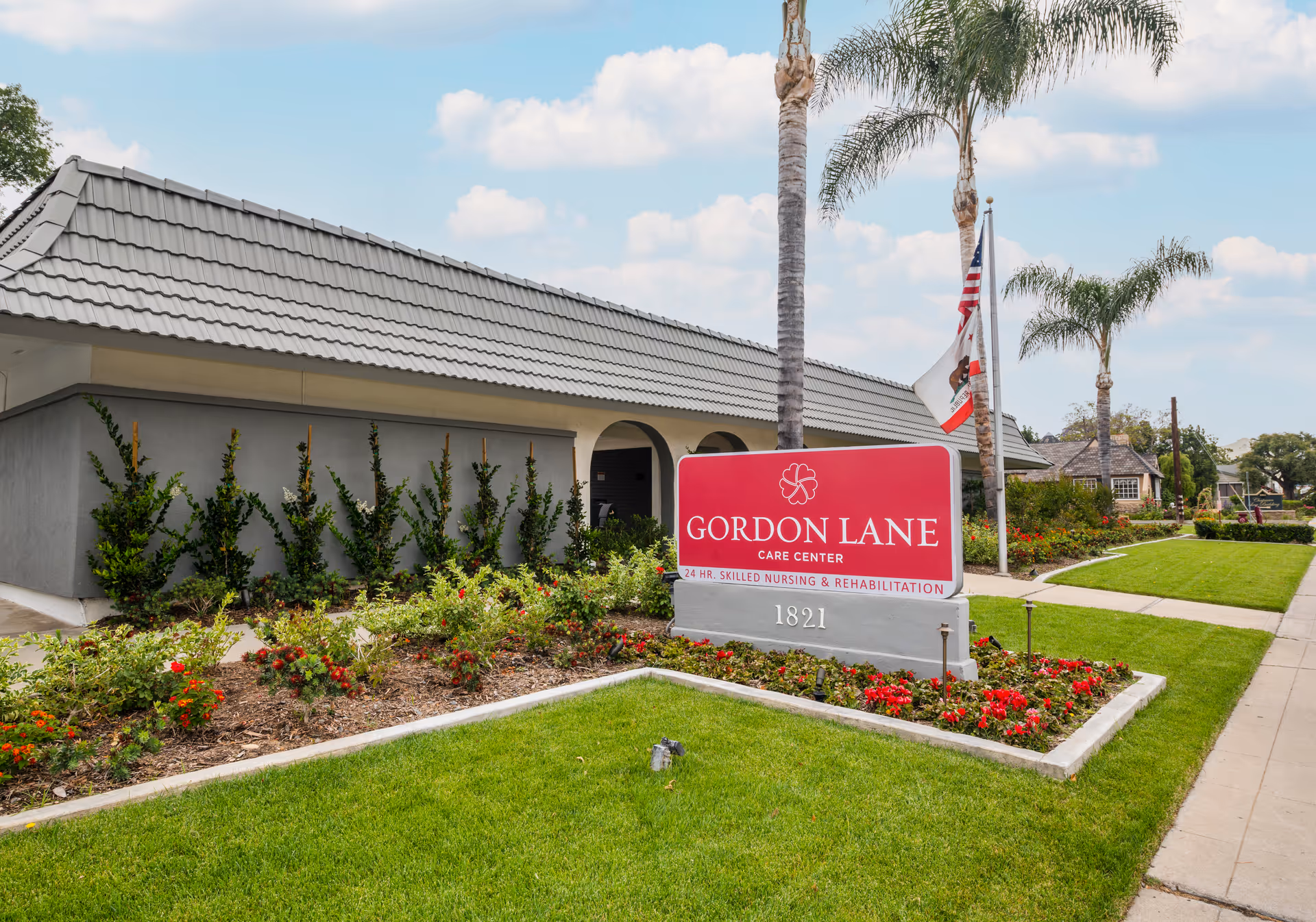 Exterior view of Gordon Lane Care Center showing a building with a tiled roof, a landscaped garden with flowers and shrubs, a sidewalk, and a sign displaying the facility's name and address. Palm trees and flags are also visible near the sign.