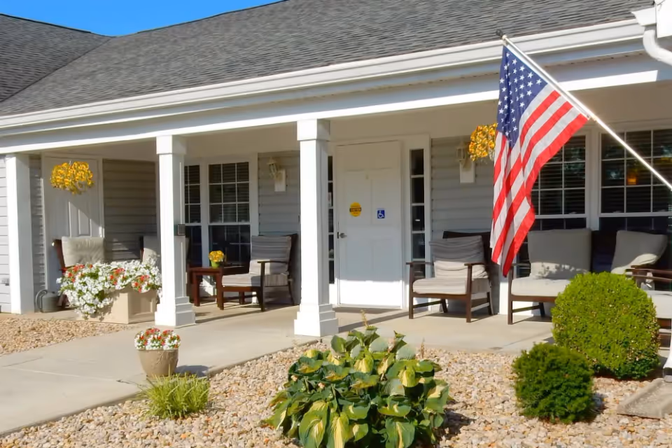 Front porch of a senior living facility with white columns, several cushioned chairs, hanging flower baskets, potted plants, and an American flag mounted near the entrance door. The building exterior is light gray with white trim and windows with blinds.