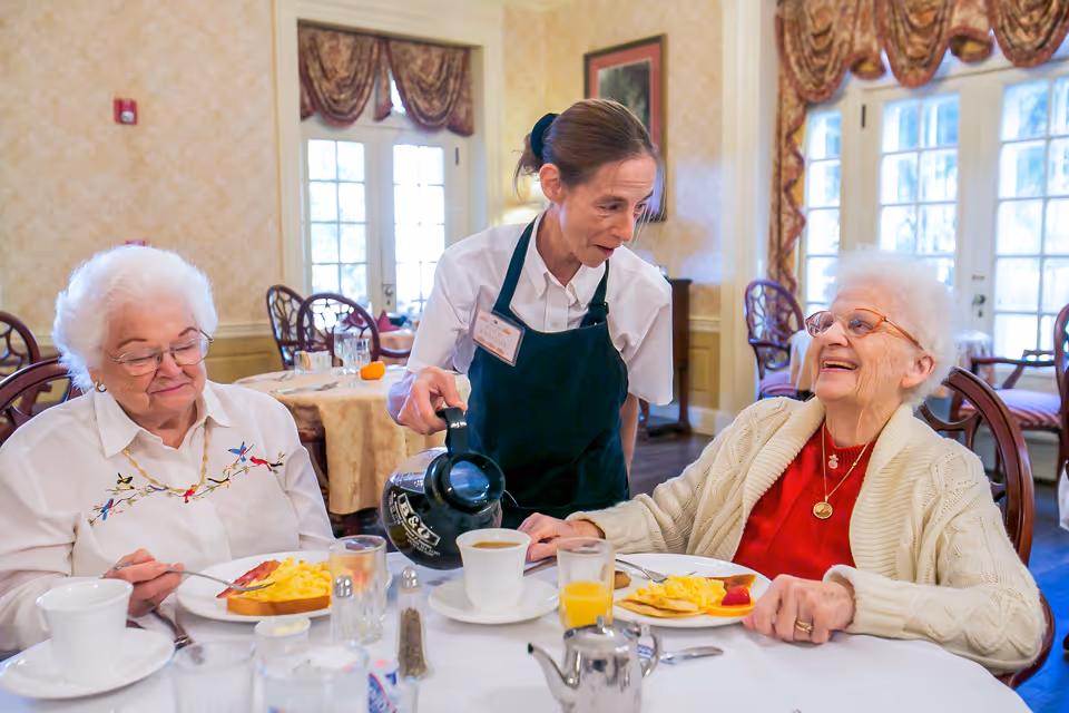 A dining room scene at Sugar Hill Assisted Living where a staff member wearing a white shirt and dark apron is pouring coffee for two elderly women seated at a table. The women are enjoying breakfast with plates of scrambled eggs, toast, and fruit. The room has large windows with draped curtains and wooden chairs.
