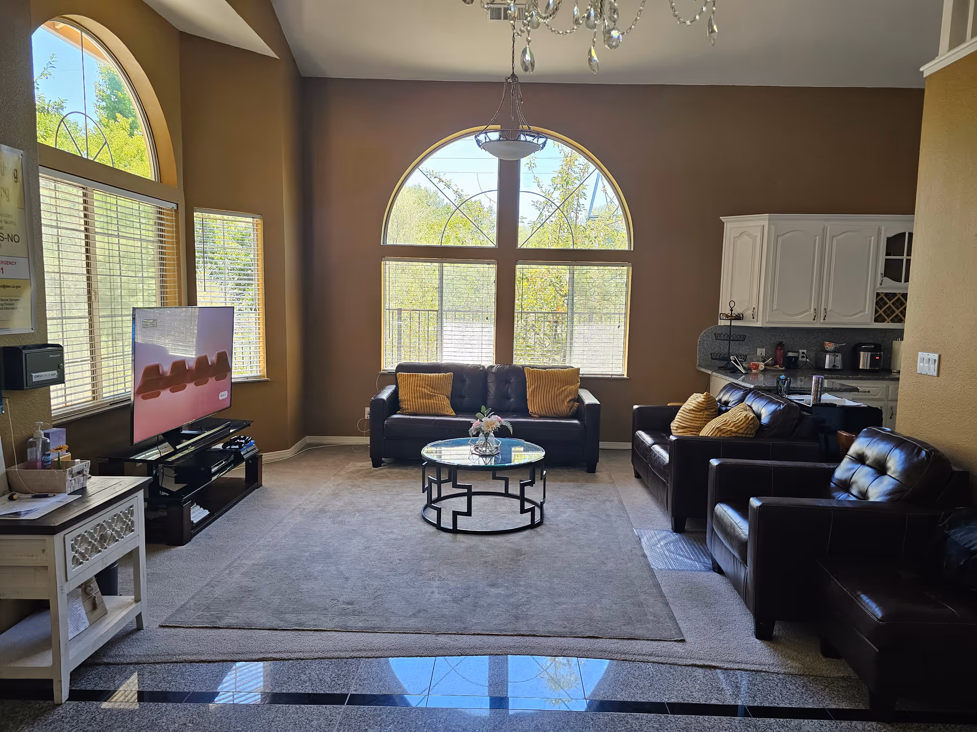 A bright living room with large arched windows letting in natural light. The room features a dark leather sofa and armchairs with yellow cushions, a round glass coffee table with a flower arrangement, a flat-screen TV on a stand, and a small white side table. The walls are painted brown, and there is a chandelier hanging from the ceiling. A kitchen area with white cabinets and a countertop is visible in the background.