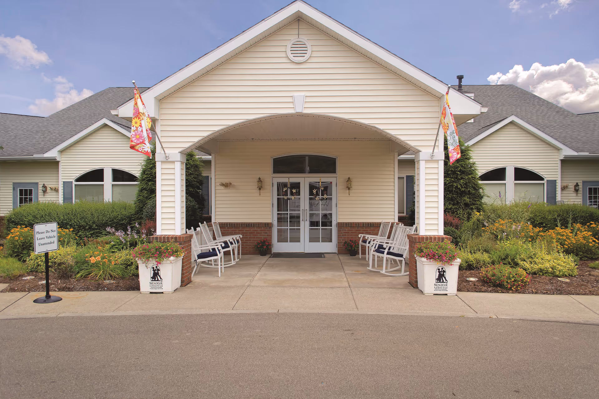 Front entrance of a senior living facility with a covered porch, white rocking chairs on either side, flower pots, and well-maintained landscaping with bushes and flowers. The building has beige siding with white trim and a gray shingled roof under a partly cloudy sky.