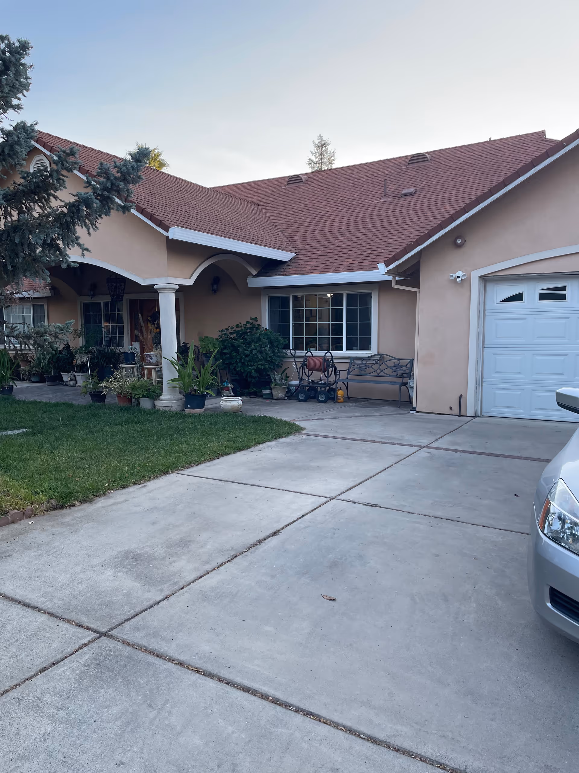 Front exterior view of a single-story house with a red shingle roof, beige walls, a white garage door, and a concrete driveway. There are several potted plants near the entrance and a bench under a window. A silver car is partially visible on the right side of the driveway.