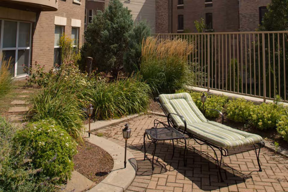 Outdoor patio area with a cushioned lounge chair and a small black metal table on a brick-paved surface, surrounded by various green plants and bushes, with a building and railing in the background.