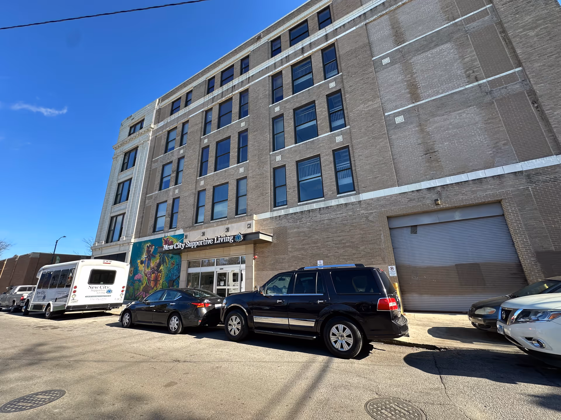 Exterior view of a multi-story brick building under a clear blue sky. The building has several windows and a sign above the entrance that reads 'New City Supportive Living.' Several cars and a shuttle bus with the same facility name are parked along the street in front of the building.