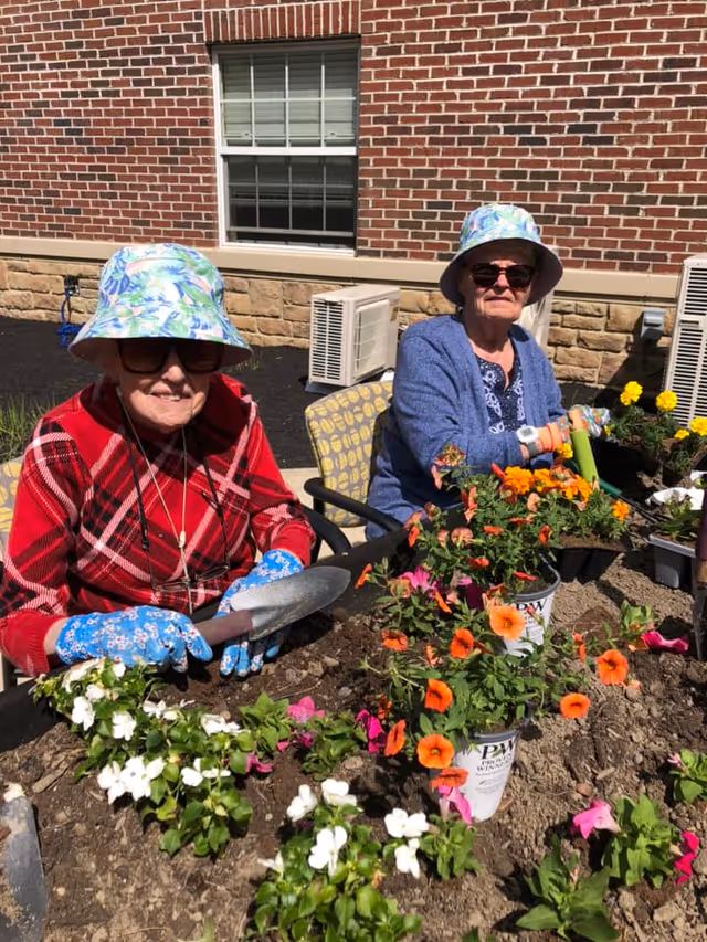 Two elderly women wearing sun hats and sunglasses are gardening outdoors in front of a brick building. They are sitting in chairs and planting colorful flowers in the soil, using gardening tools. One woman is wearing a red plaid shirt and blue floral gloves, while the other is wearing a blue cardigan and multiple bracelets.