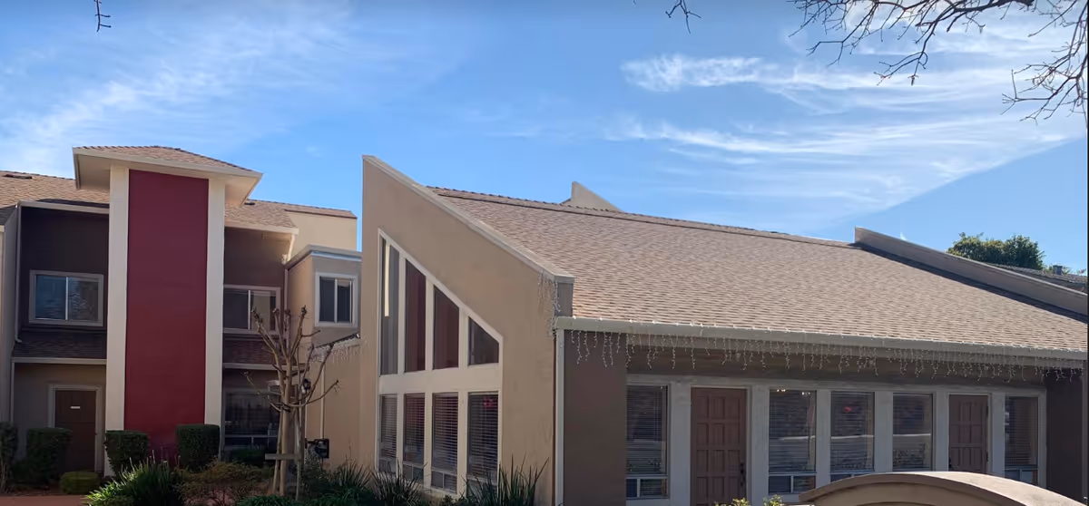 Exterior view of a residential building with a sloped roof, multiple windows, and doors. The building has beige and brown walls with a red vertical accent. There are some bushes and a small tree in front of the building under a blue sky with light clouds.