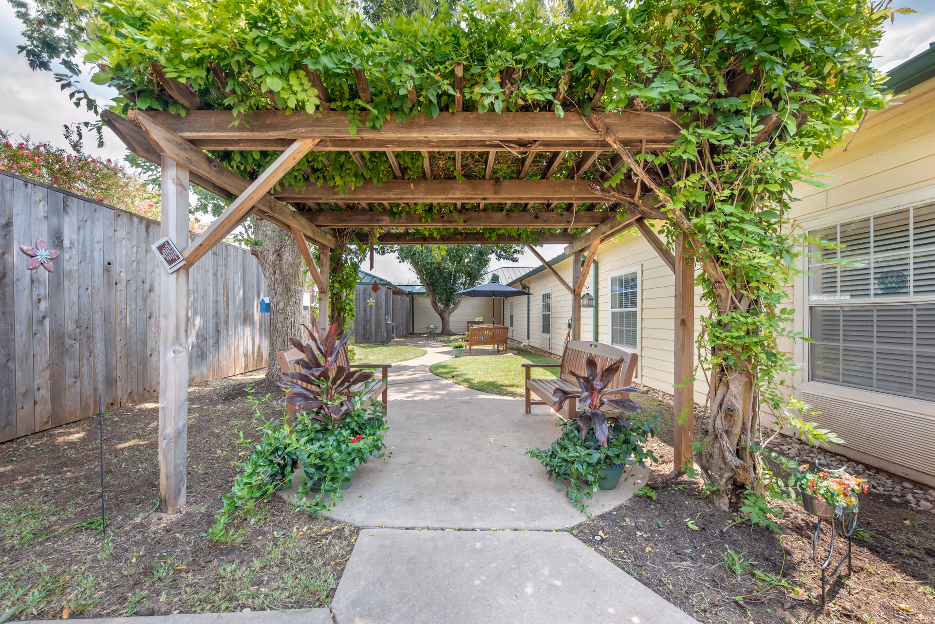 Outdoor garden area at Brookdale Beckett Meadows featuring a wooden pergola covered with green vines. There are two wooden benches with potted plants on either side of the concrete pathway. The pathway leads to a seating area with a bench and umbrella, surrounded by grass, trees, and a wooden fence.