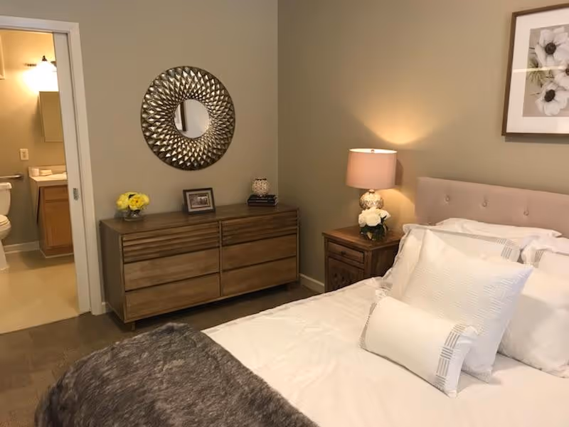 A cozy bedroom with a neatly made bed featuring white pillows and a gray throw blanket. To the right of the bed is a wooden nightstand with a lamp and a small flower arrangement. On the wall above the nightstand is a framed floral artwork. Across from the bed is a wooden dresser with decorative items including a round metallic mirror, a small framed photo, a candle, and a vase with yellow flowers. An open door reveals a bathroom with a sink, mirror, and toilet.