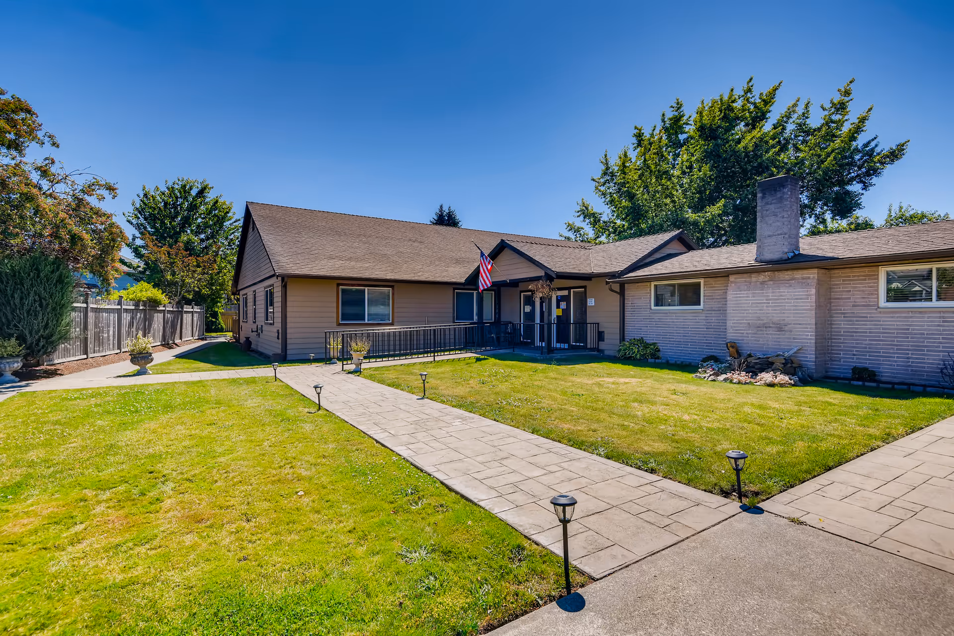 Exterior view of a single-story building with a brown roof and beige siding, surrounded by a well-maintained lawn and paved walkways. An American flag is displayed near the entrance, which has a ramp and handrails. Trees and a wooden fence are visible in the background under a clear blue sky.