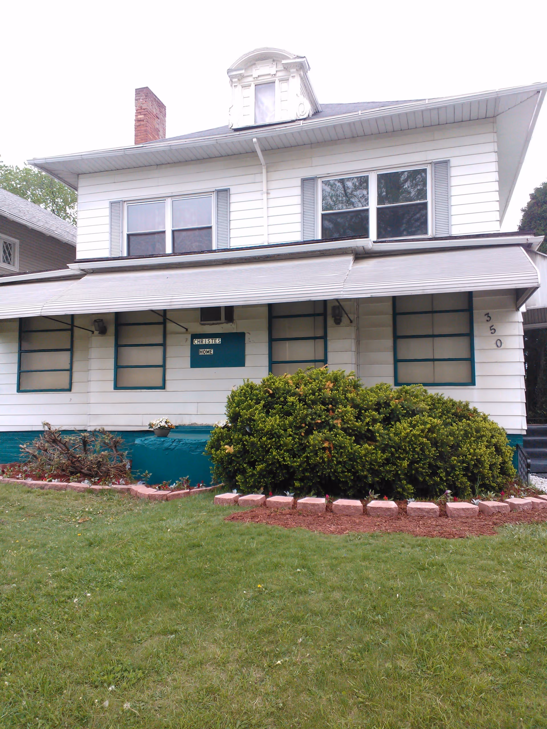Exterior view of a two-story white house with blue trim and a large awning over the front porch. The house has multiple windows, a chimney, and a small dormer window on the roof. There is a green lawn and landscaped bushes in front of the house. A sign on the porch reads 'Christies Home' and the house number 350 is visible on the right side.