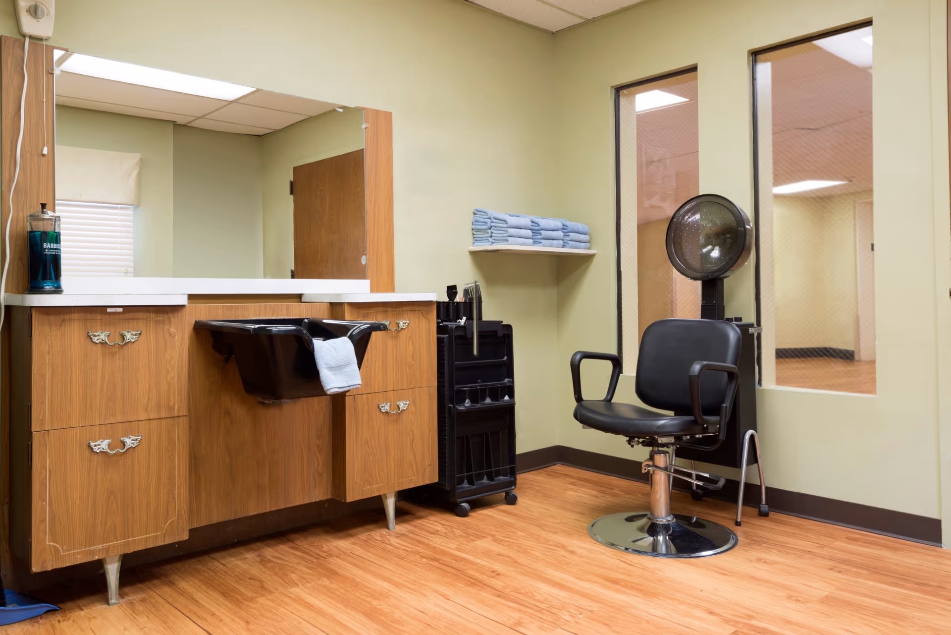 Interior of a small hair salon area with a wooden cabinet featuring a black wash basin, a large mirror above the cabinet, a black salon chair with armrests, a hair dryer hood, a shelf with neatly folded blue towels, and a rolling cart with hair styling tools. The room has light green walls and wood-patterned flooring.