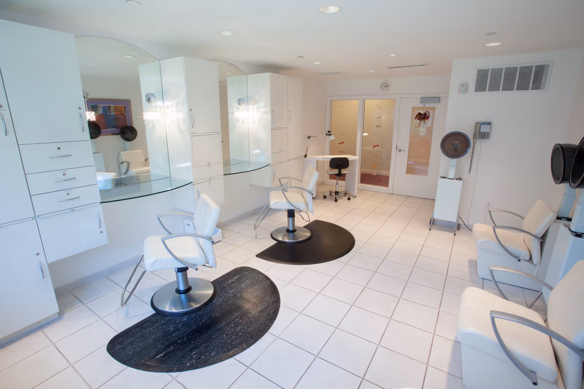 A clean and modern salon area with white swivel chairs on black mats, large mirrors mounted on the wall, white cabinets, and hair drying stations. The room has white tiled flooring and bright overhead lighting.