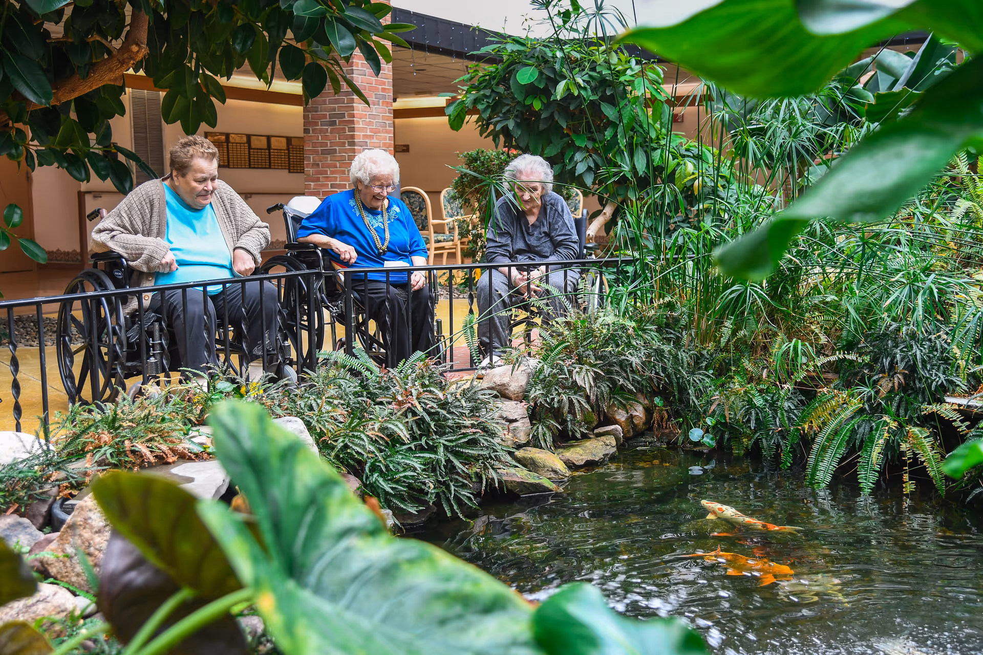 Three elderly women, two in wheelchairs and one sitting, are gathered around an indoor pond with koi fish. The area is lush with green plants and has a railing around the pond. The setting appears to be a peaceful indoor garden or atrium within a senior living facility.