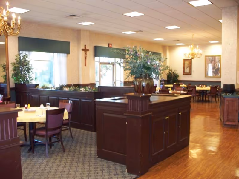 Interior view of a dining room in a senior living facility with multiple tables and chairs arranged for meals. The room features wooden furniture, a large floral arrangement on a central wooden cabinet, chandeliers, framed artwork on the walls, and windows with green valances allowing natural light to enter.