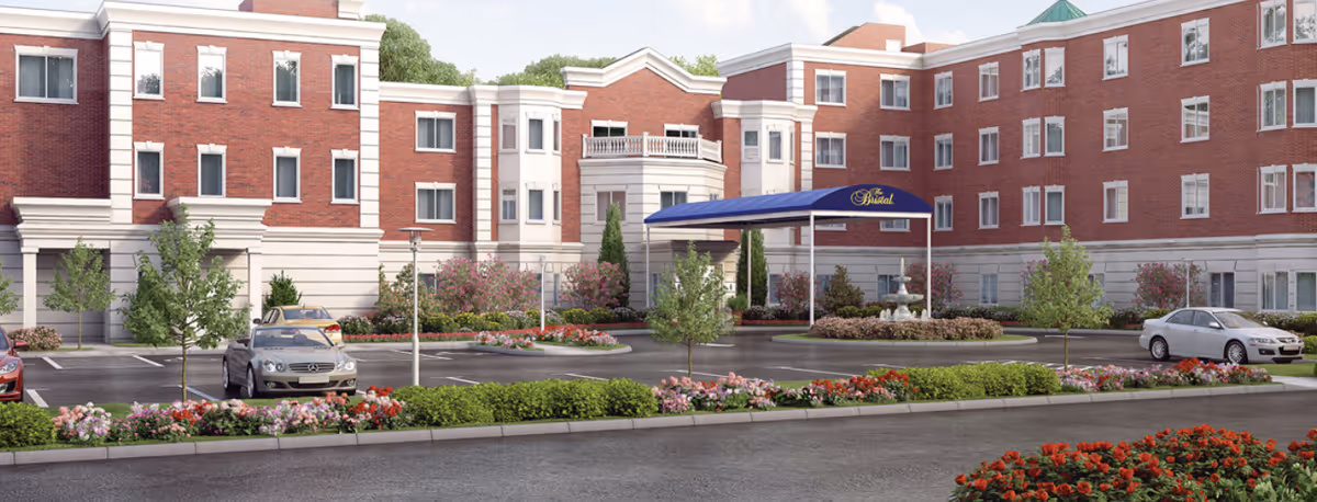 Exterior view of The Bristal Assisted Living at Holtsville building with a circular driveway, landscaped flower beds, small trees, and parked cars. The building is made of red brick with white trim and has multiple windows and a blue canopy over the entrance.