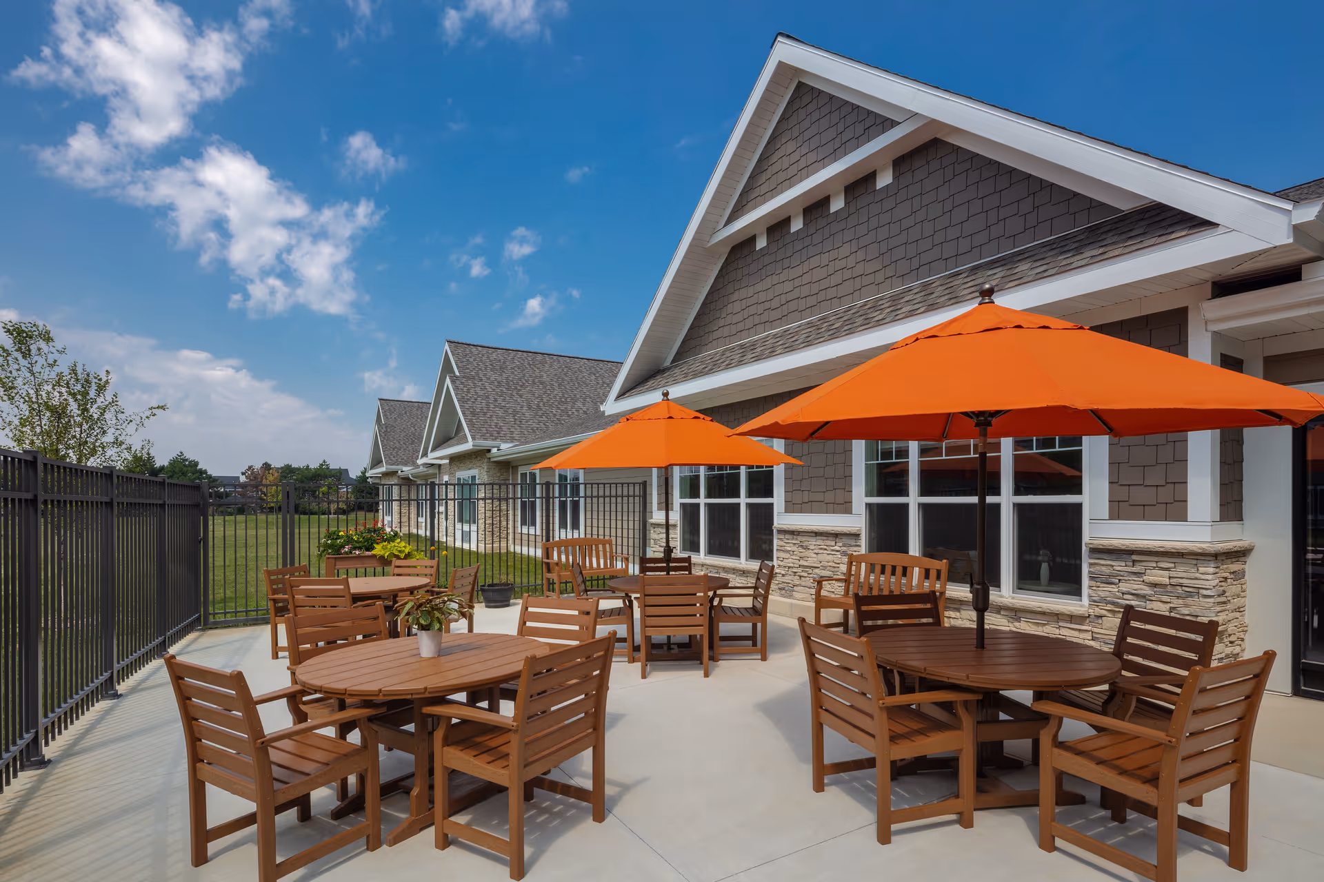 Outdoor patio area at The Waters of Pewaukee featuring multiple round wooden tables with matching chairs and bright orange umbrellas. The patio is adjacent to a building with gray siding and stone accents, under a blue sky with scattered clouds.