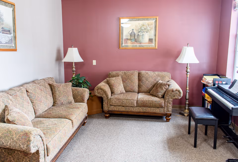A cozy living room with two patterned sofas facing each other, two floor lamps with white shades, a framed painting on a maroon accent wall, a potted plant, and an electronic keyboard with a bench near a window.
