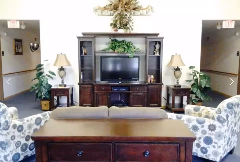 A living room area with a wooden entertainment center holding a flat-screen TV, flanked by two table lamps on side tables and two patterned armchairs facing the TV. There are plants on either side of the entertainment center and hallways visible in the background.