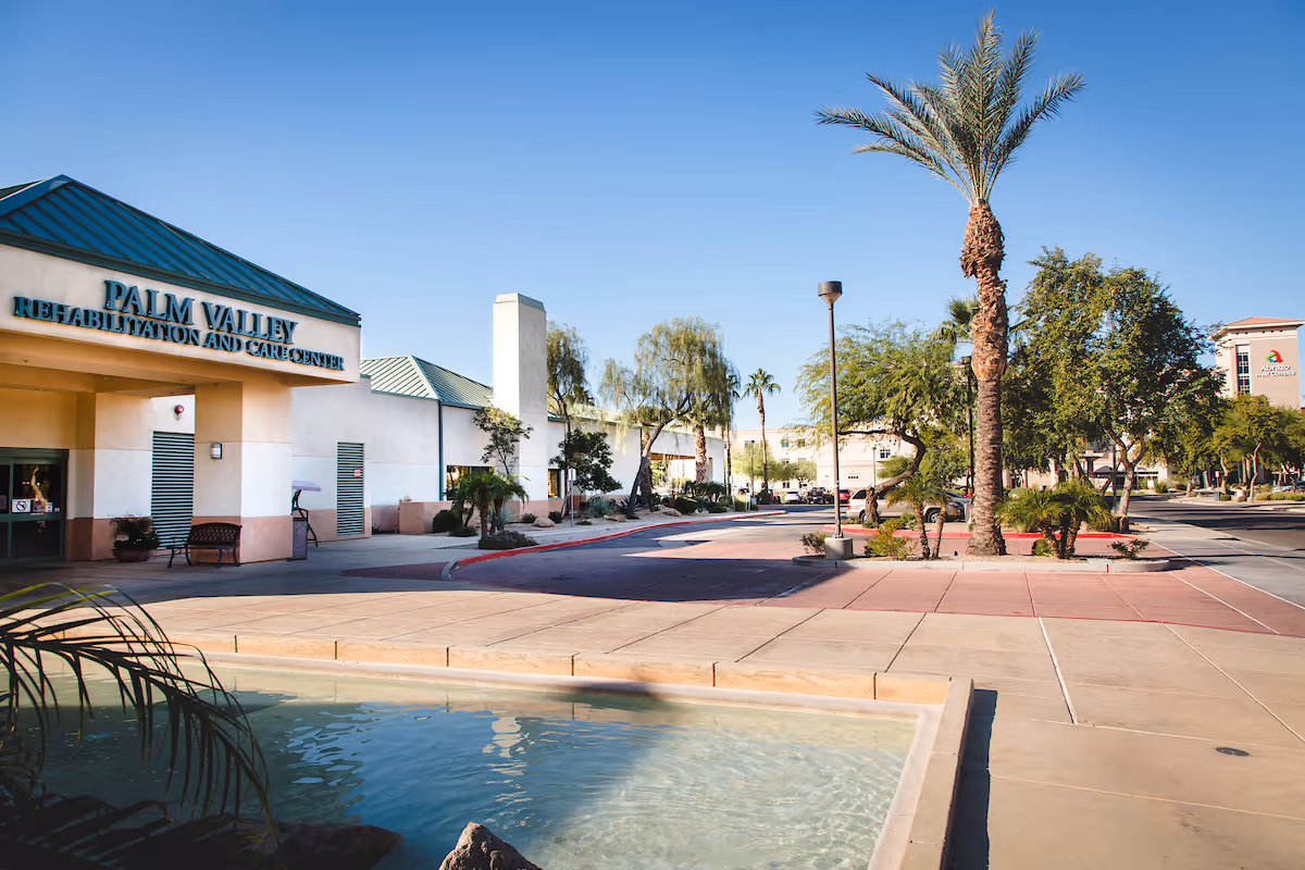 Exterior view of Palm Valley Rehabilitation and Care Center building with a small water feature in the foreground, palm trees, and a clear blue sky.