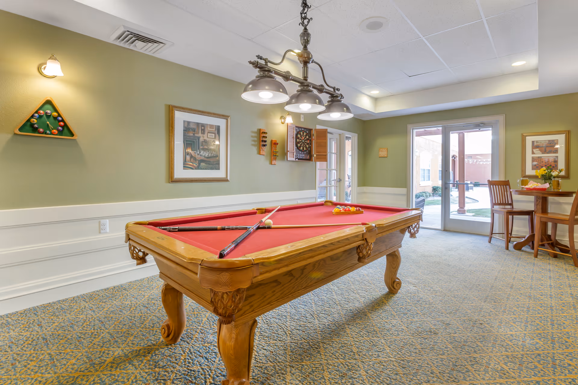 A recreational room with a red felt pool table in the center, two pool cues resting on it, and a rack of billiard balls on the table. The room has green walls with white wainscoting, a dartboard mounted on the wall, framed artwork, and a triangular wall-mounted rack holding billiard balls. There is a hanging light fixture above the pool table and a small round table with two wooden chairs near glass doors leading outside to a patio area.