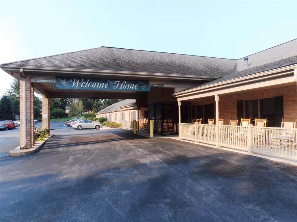 Entrance of Evergreen Estates Retirement Community with a covered driveway and a sign that reads 'Welcome Home'. There are parked cars in the background and a porch area with several rocking chairs on the right side.