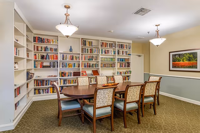 A cozy library or reading room with a large wooden table surrounded by eight cushioned chairs. The walls are lined with built-in white bookshelves filled with books. Two ceiling light fixtures hang above the table, and a framed landscape photograph is on the wall. The room has a carpeted floor and soft, neutral-colored walls.