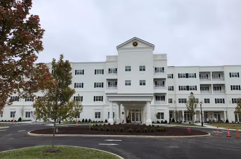 Front exterior view of a large, white senior living facility building with multiple windows, balconies, and a central entrance with columns. There is a circular driveway with landscaped areas and young trees in front of the building under a cloudy sky.