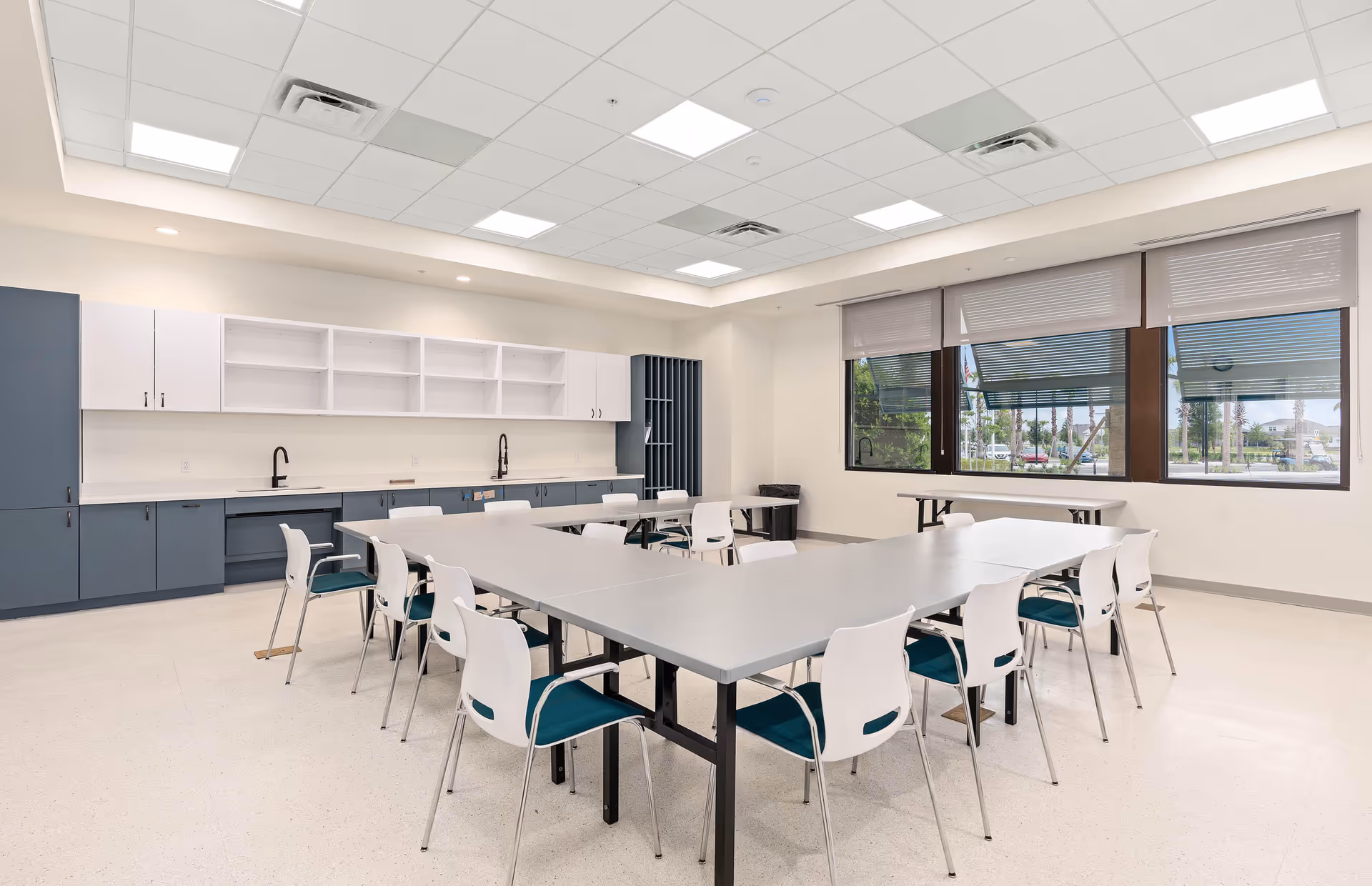 A bright, modern multipurpose room with large windows, white walls, and a ceiling with recessed lighting. The room features a large U-shaped arrangement of gray tables surrounded by white chairs with teal cushions. Along one wall, there are blue and white cabinets with two black faucets and sinks. The windows have partially lowered blinds and offer a view of the outdoor area.